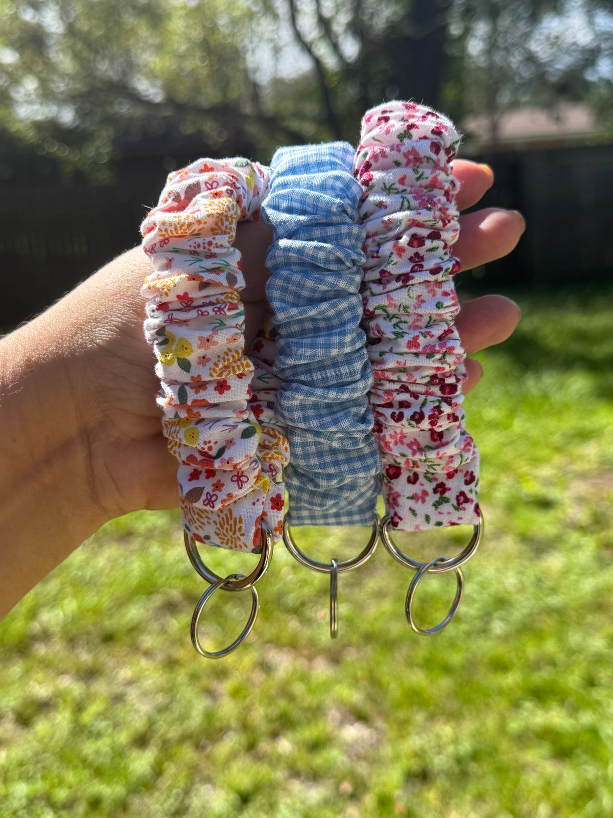 Three fabric key rings held in a hand outdoors, featuring floral and gingham patterns in pink, blue, and white, with metal key rings at the bottom.