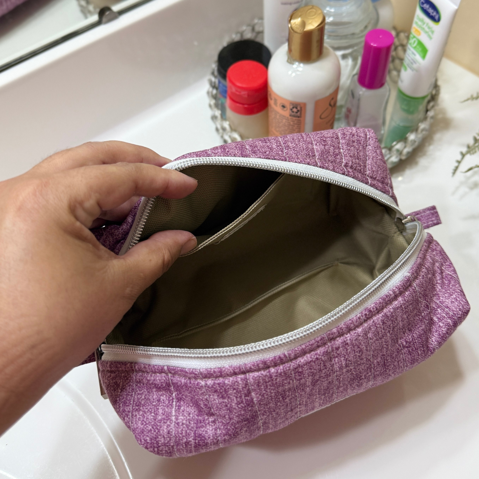 A lavender purple textured cosmetic pouch with a white zipper, held open by a hand over a white bathroom sink with toiletries in the background.