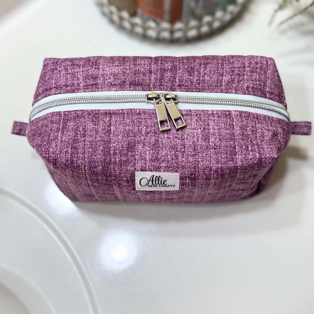 A textured purple fabric cosmetic pouch with a white zipper and a small white brand label reading 'Allie's Handmade', resting on a glossy white surface.