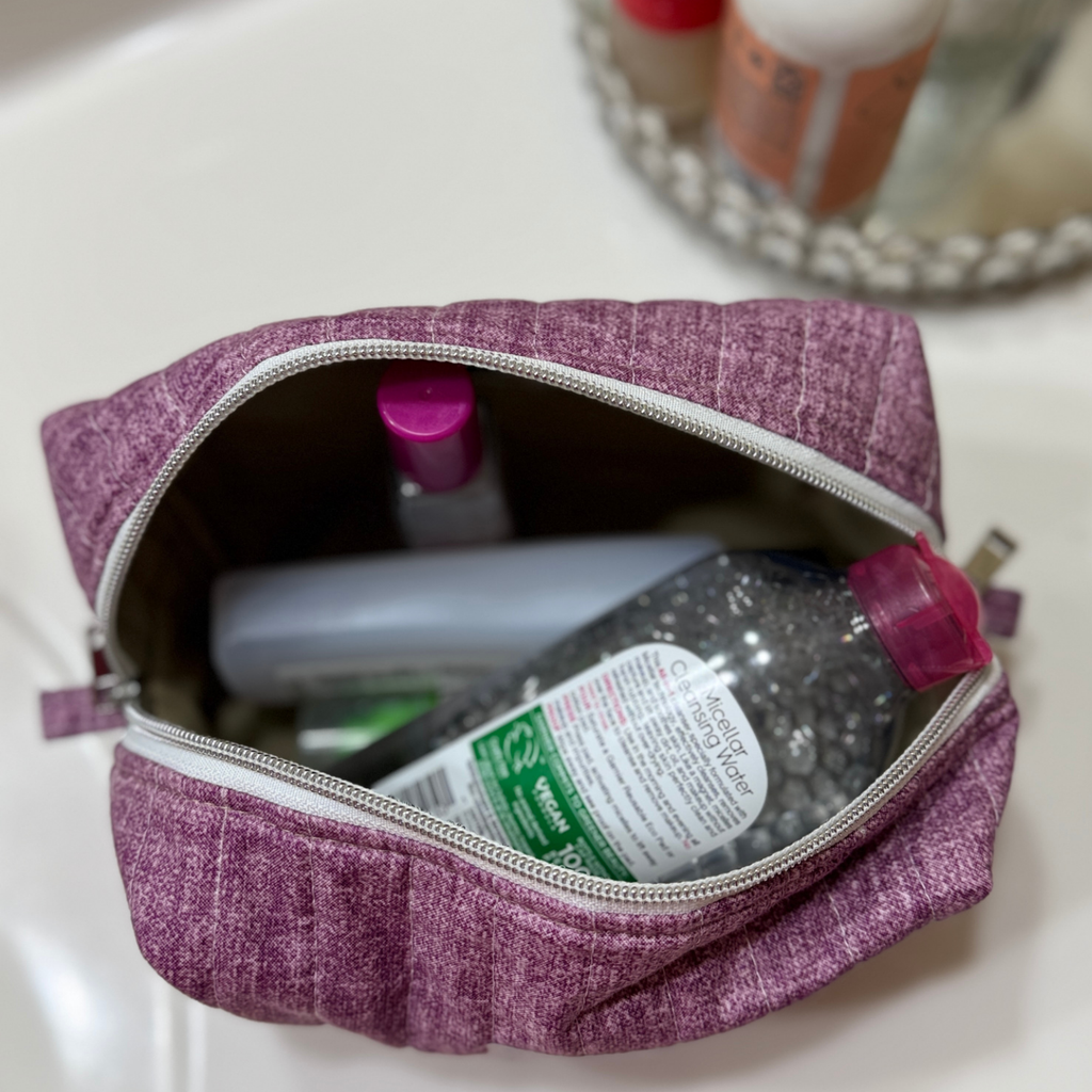 A purple textured fabric makeup bag with a silver zipper, shown open on a white sink surface with toiletries inside.