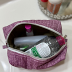 A purple textured fabric makeup bag with a silver zipper, shown open on a white sink surface with toiletries inside.