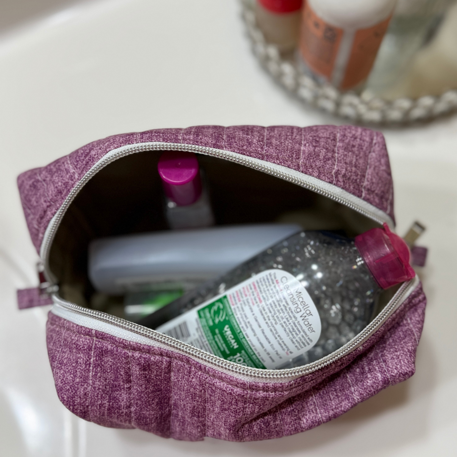 A purple textured fabric makeup bag with a silver zipper, shown open on a white sink surface with toiletries inside.