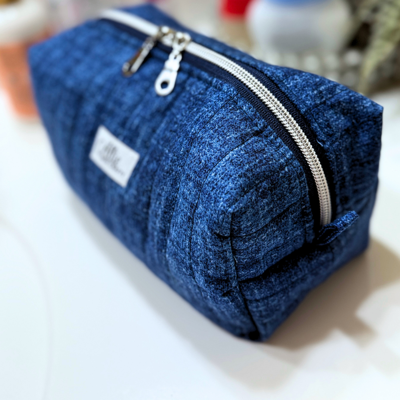A blue quilted fabric zippered pouch with a silver zipper and a small white label, photographed on a white surface with a softly blurred background.