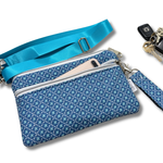A blue patterned crossbody pouch with a matching blue wrist strap and a zippered pocket holding a phone, shown on a white background with keys nearby.
