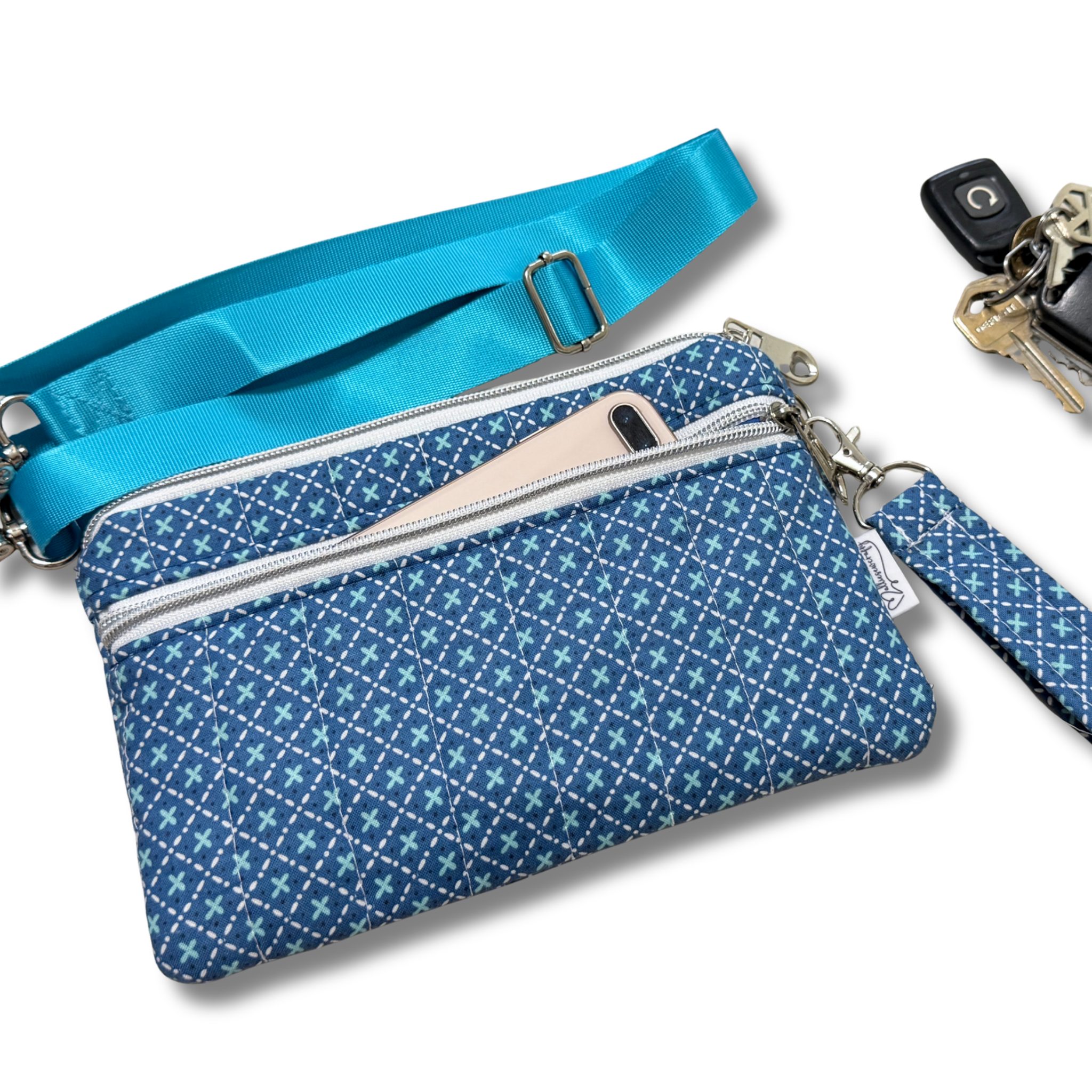 A blue patterned crossbody pouch with a matching blue wrist strap and a zippered pocket holding a phone, shown on a white background with keys nearby.