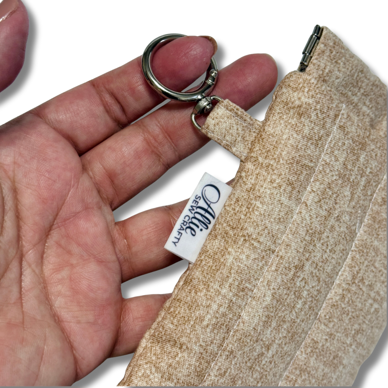 Hand holding a cream pouch by the round swivel hook in a white background with Allie Sew Crafty label