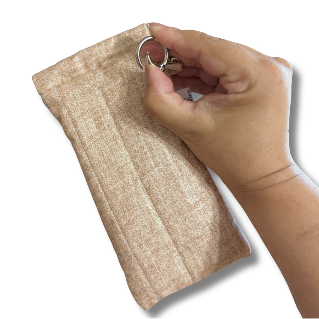 A close-up of a hand holding a beige textured fabric pouch with a silver metal ring attachment, shown against a plain white background.