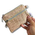 A small zippered coin pouch in a textured beige fabric, held by a hand against a plain white background. The pouch features a silver zipper, a small metal ring, and a white fabric tag with the brand name 'Allie Sew Crafty'.