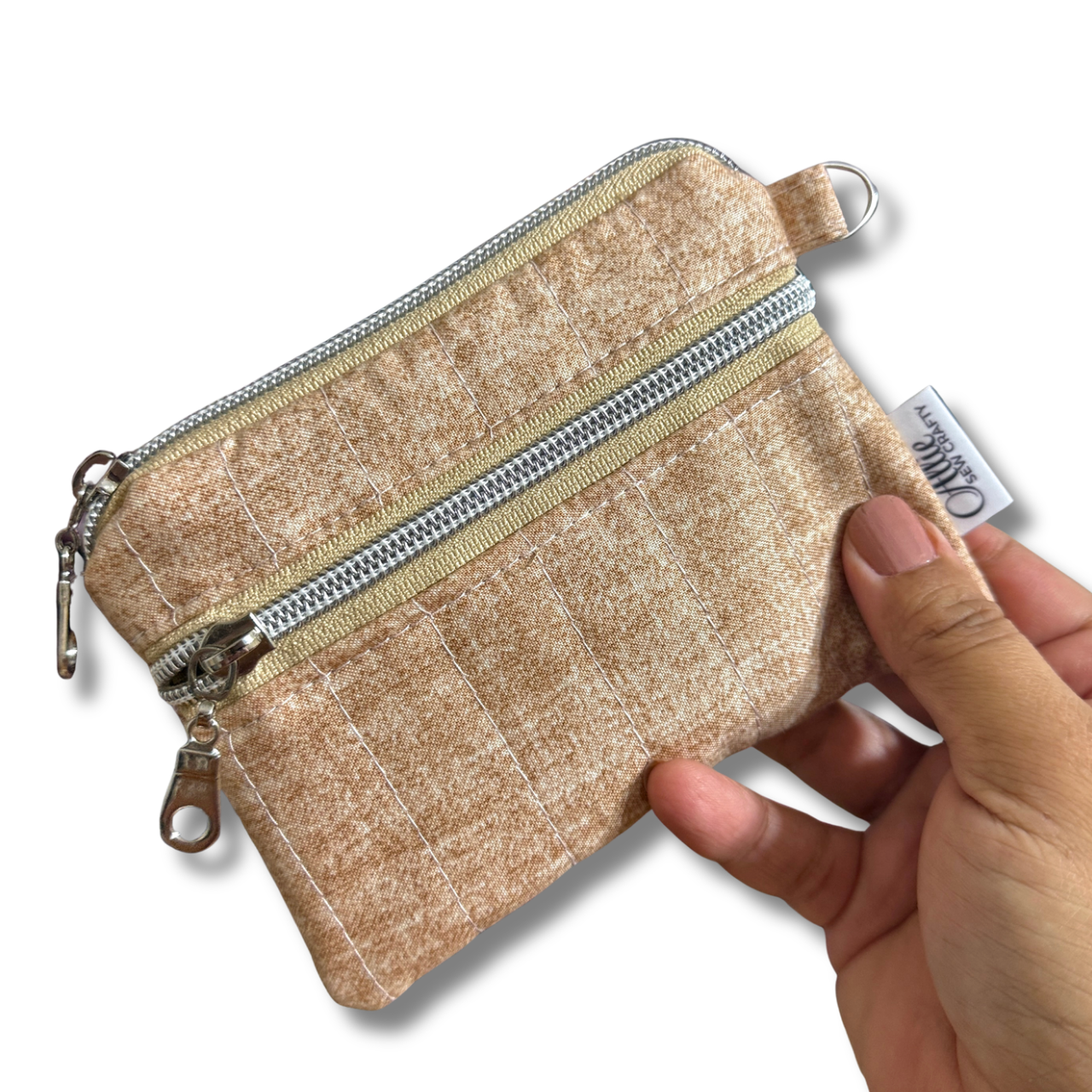 A small zippered coin pouch in a textured beige fabric, held by a hand against a plain white background. The pouch features a silver zipper, a small metal ring, and a white fabric tag with the brand name 'Allie Sew Crafty'.