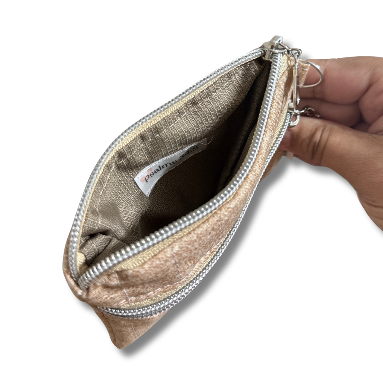 A close-up of a metallic beige zippered pouch being held open by a hand, showing the interior lining and a brand tag that reads 'Psalms 34:5.