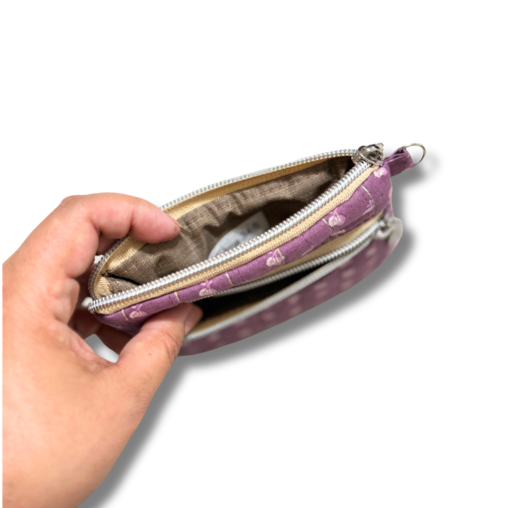 A hand holding open a small zippered pouch with a lavender purple fabric exterior decorated with small white hearts, showing a beige lining inside.