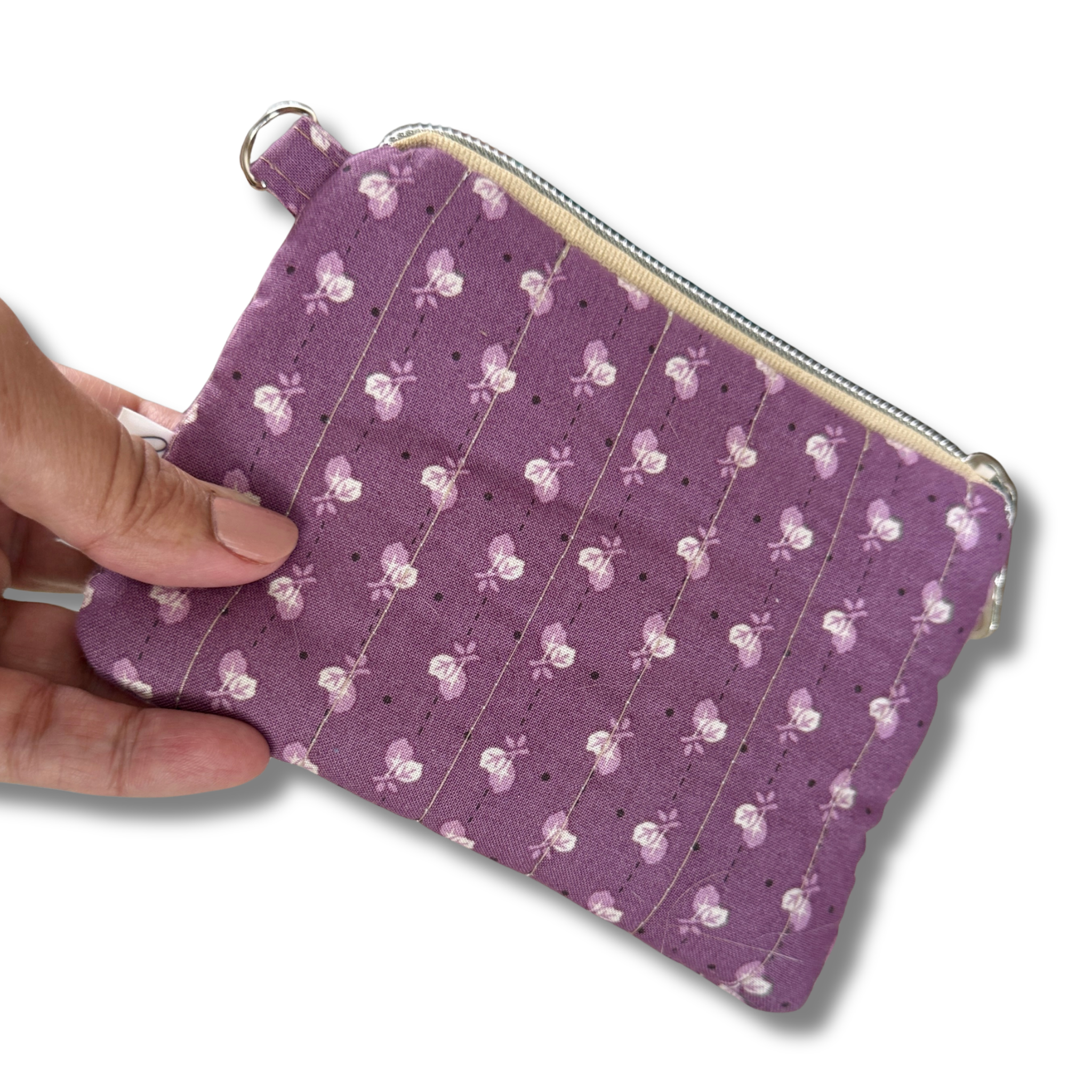 A hand holding a small quilted fabric pouch in lavender purple with a white and pink floral pattern, shown against a plain white background.