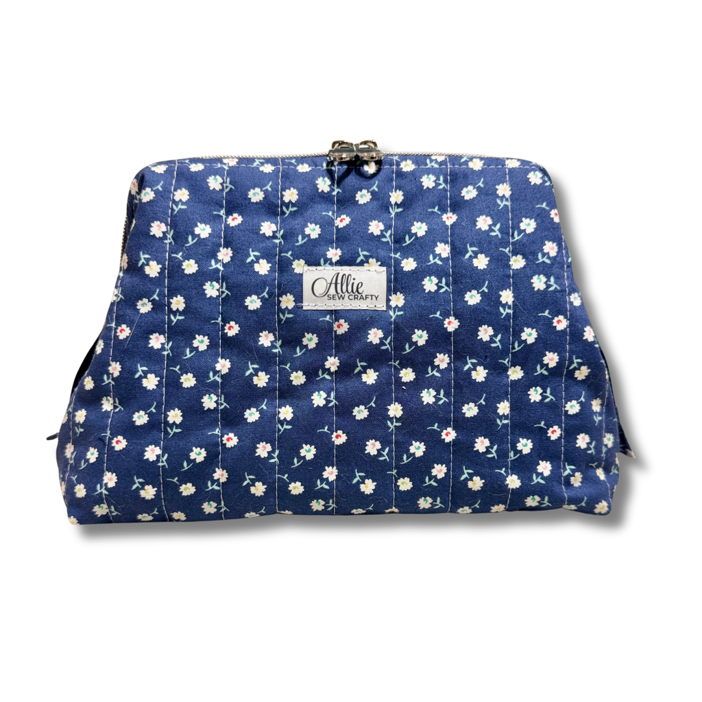 A quilted cosmetic pouch by Allie's Handbag featuring a navy blue background with a white and pink floral vine pattern, a silver-tone metal clasp at the top, and a white brand label on the front.