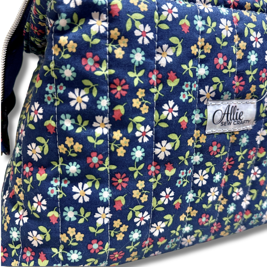 close up of quilting cotton fabric navy blue background with a colorful floral pattern of pink, yellow, white, and green flowers and Allie Sew Crafty in a white label.