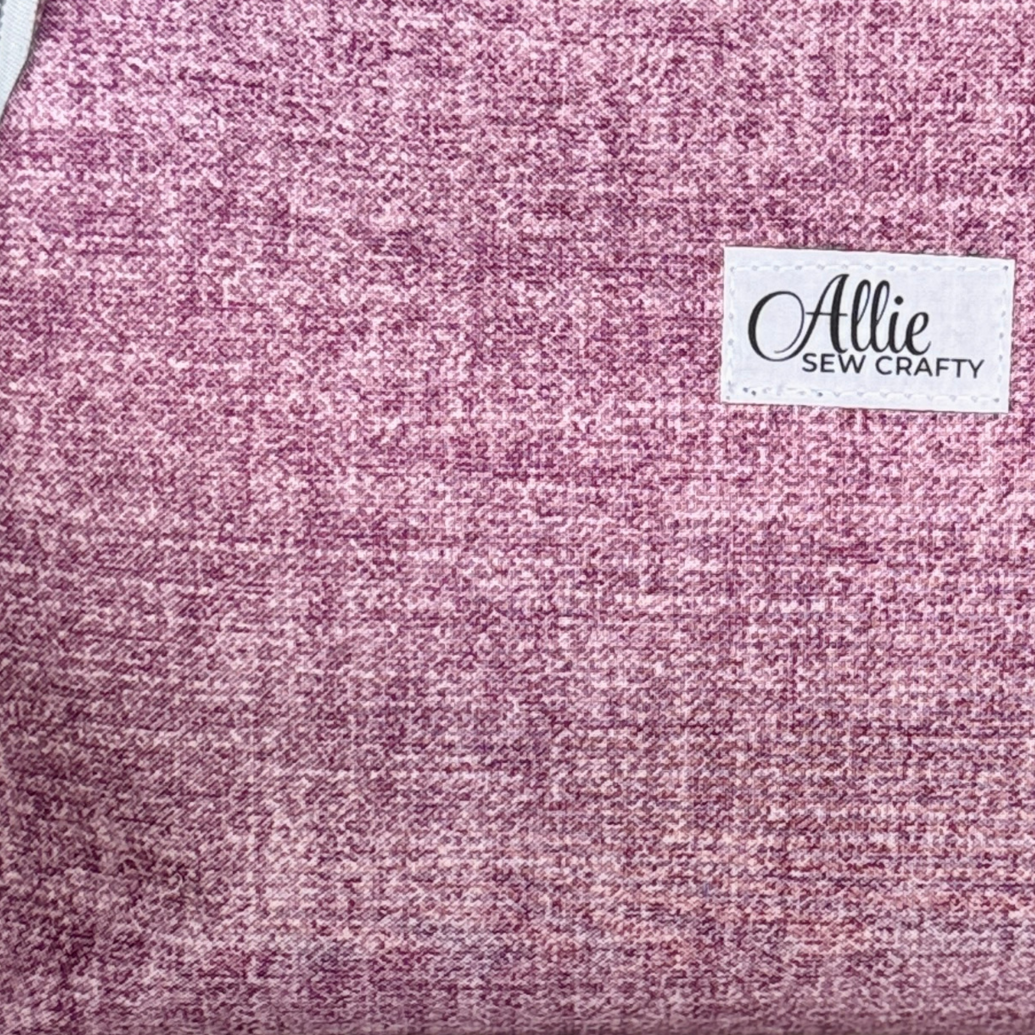 A close-up of a textured fabric in a heathered pink and purple tone, featuring a white brand label that reads 'Allie Sew Crafty'.
