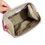 An open cosmetic pouch or makeup bag shown from the inside, with a hand holding the left side to display the interior. The pouch has a beige/tan interior lining, a silver zipper, and a pink patterned exterior visible at the edges.