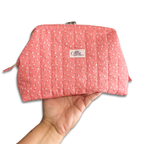 A pink quilted cosmetic pouch with a white floral pattern, held in a hand against a white background. The pouch features a central label that reads 'Allie Sew Crafty'
