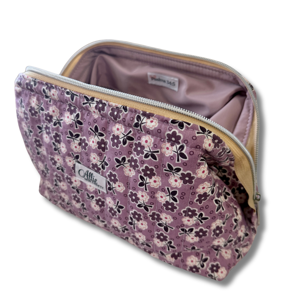 A lavender quilted cosmetic pouch with a floral print, shown open to reveal a light purple interior. The pouch features a purple, white, and black floral pattern, a silver zipper, and a brand label reading 'Allie'.