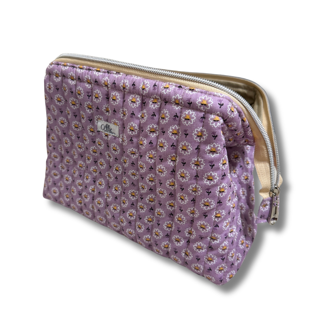 A quilted lavender cosmetic pouch with a daisy floral pattern, featuring a cream zipper and a small white label 'Allie Sew Crafty' brand label.