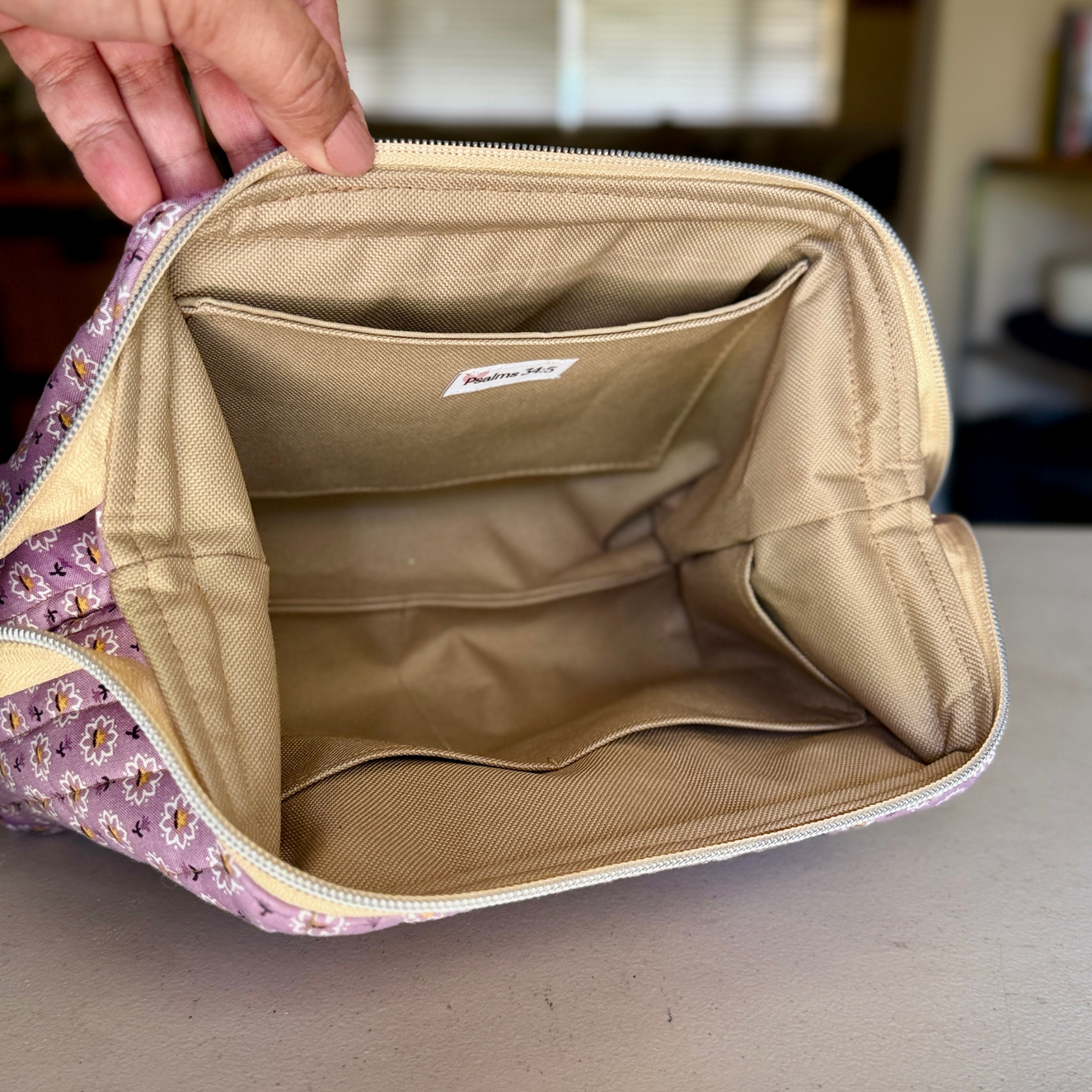 A close-up of a zippered cosmetic pouch being held open by a hand, showing a beige interior with multiple compartments and a small interior label. The exterior fabric has a lavender floral pattern.