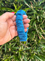 A hand holding a bright blue ruched fabric strap or keychain loop with silver metal rings, set against a background of green foliage.