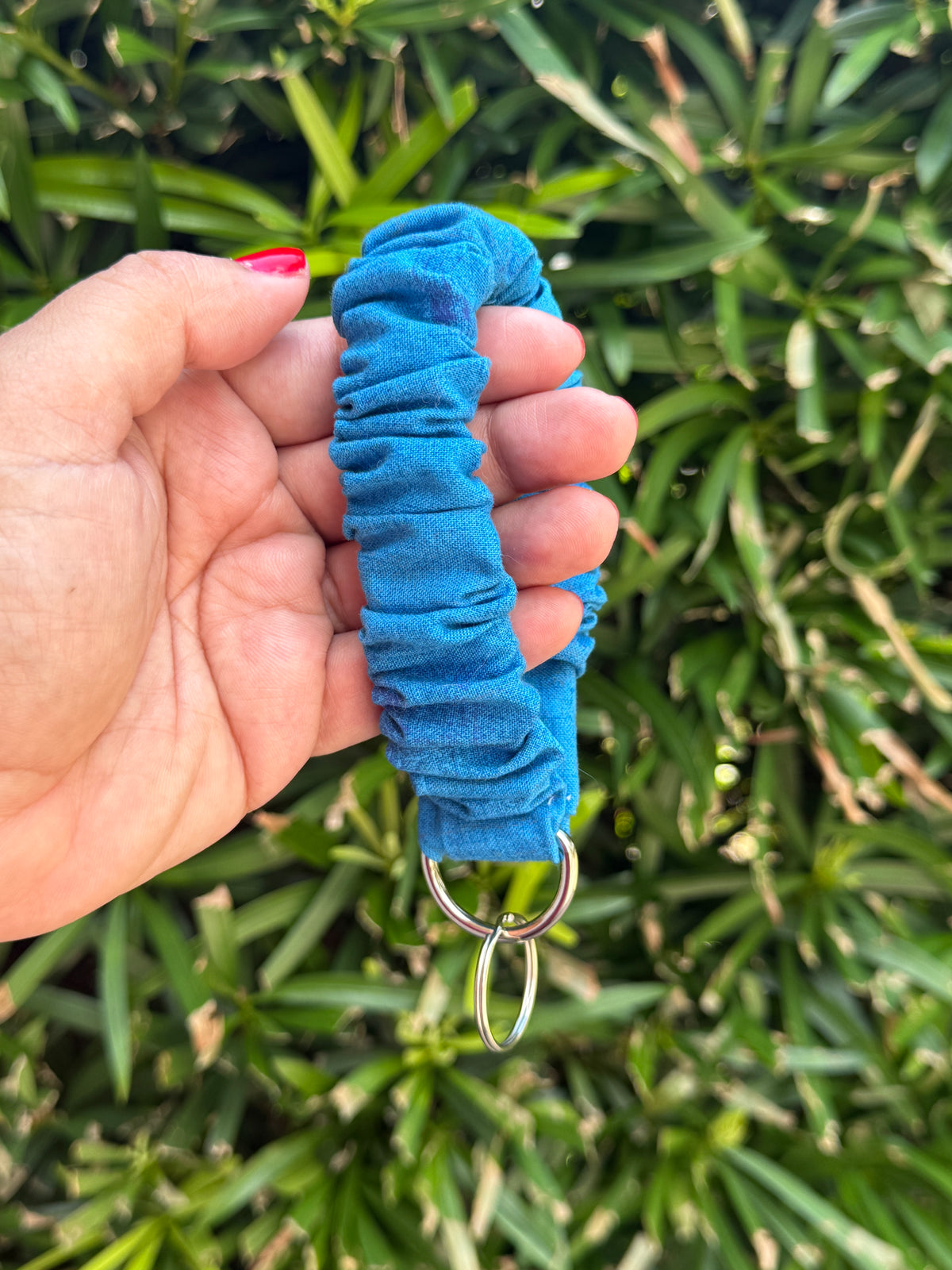 A hand holding a bright blue ruched fabric strap or keychain loop with silver metal rings, set against a background of green foliage.