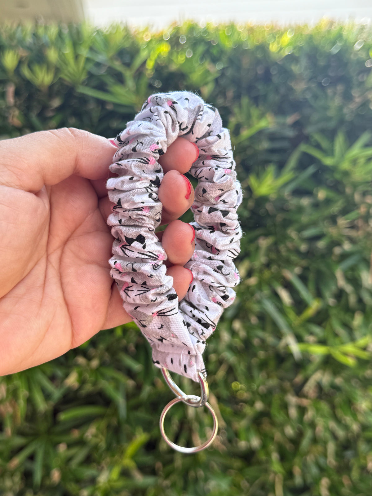 A hand holding a fabric wristlet keychain with a black and white patterned print, featuring a silver metal split ring, set against a blurred green outdoor background.