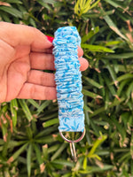 A hand holding a light blue fabric wristlet keychain with a blue floral pattern and a silver metal key ring, set against a background of green foliage.