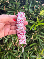 A hand with red nail polish holds a pink and red cherry-patterned fabric wrist strap keychain with a silver split ring against a background of green foliage.