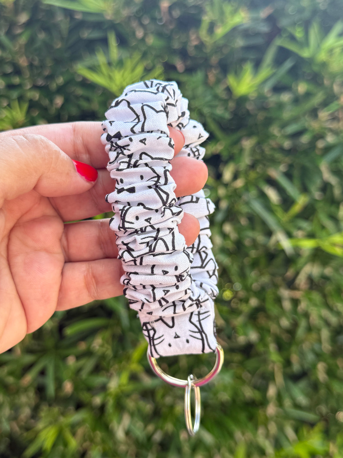 A hand holding a white fabric wristlet keychain with a black abstract line pattern and a silver metal key ring, set against a blurred green foliage background.