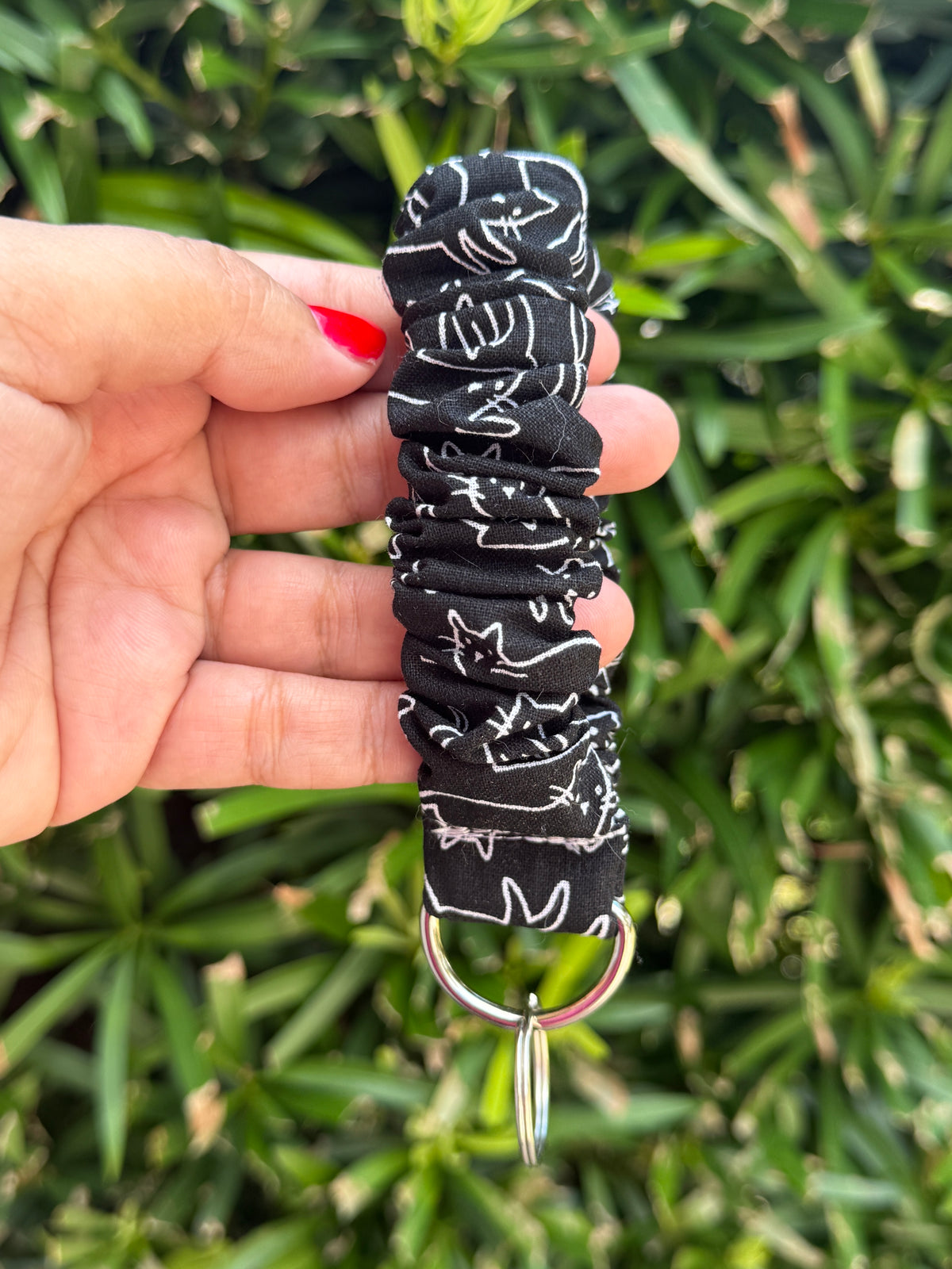 A hand holding a black fabric wristlet keychain with a white cat pattern and a silver metal key ring, set against a blurred green foliage background.