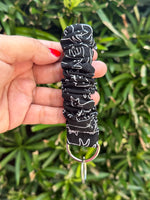 A hand holding a black fabric wristlet keychain with a white cat pattern and a silver metal key ring, set against a blurred green foliage background.