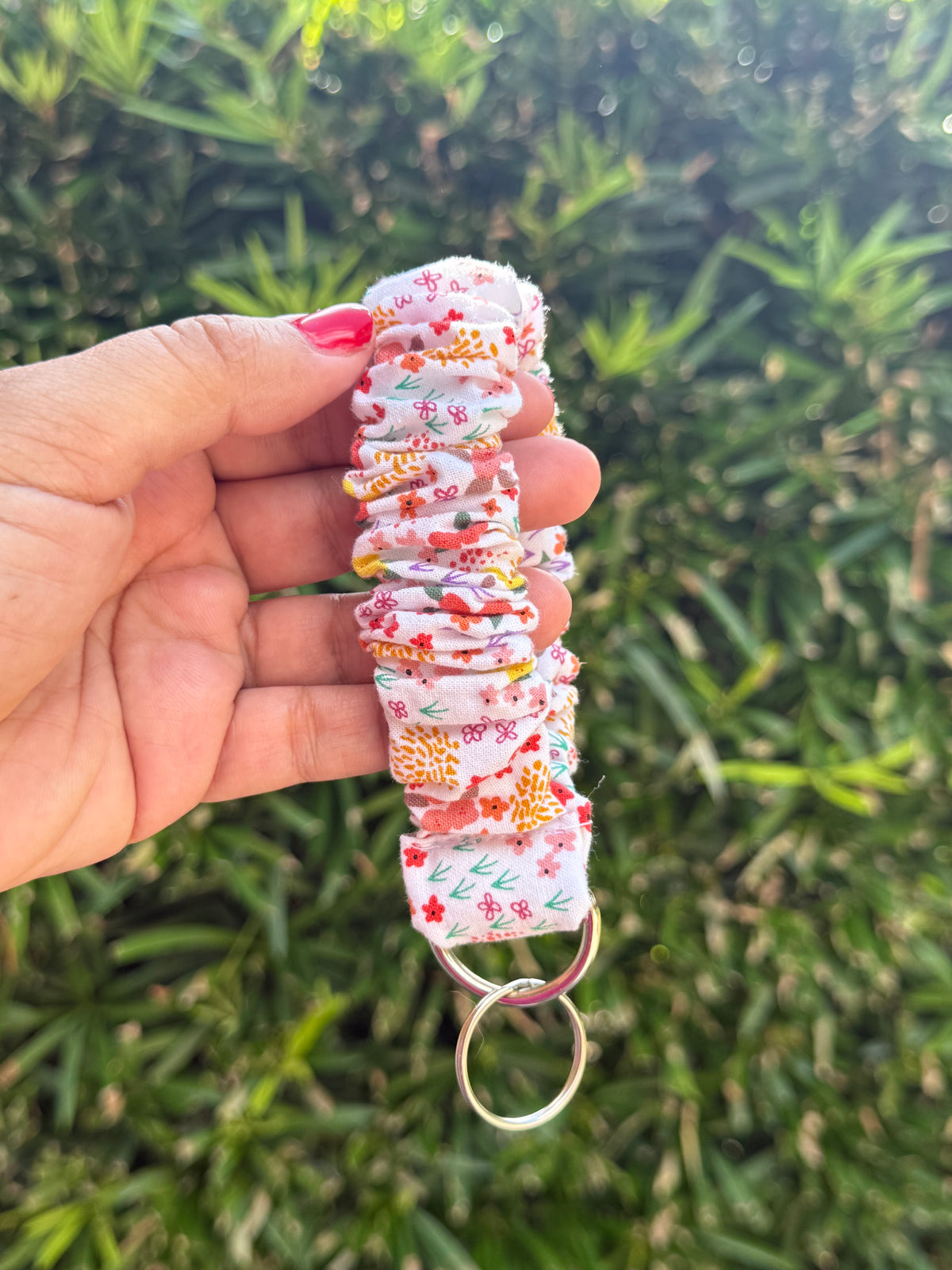 A fabric wrist strap keychain with a floral print, held by a hand against a blurred green outdoor background.