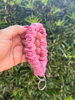 A hand holding a pink ruched fabric wristlet or keychain strap with a silver metal key ring, set against a blurred green outdoor background.