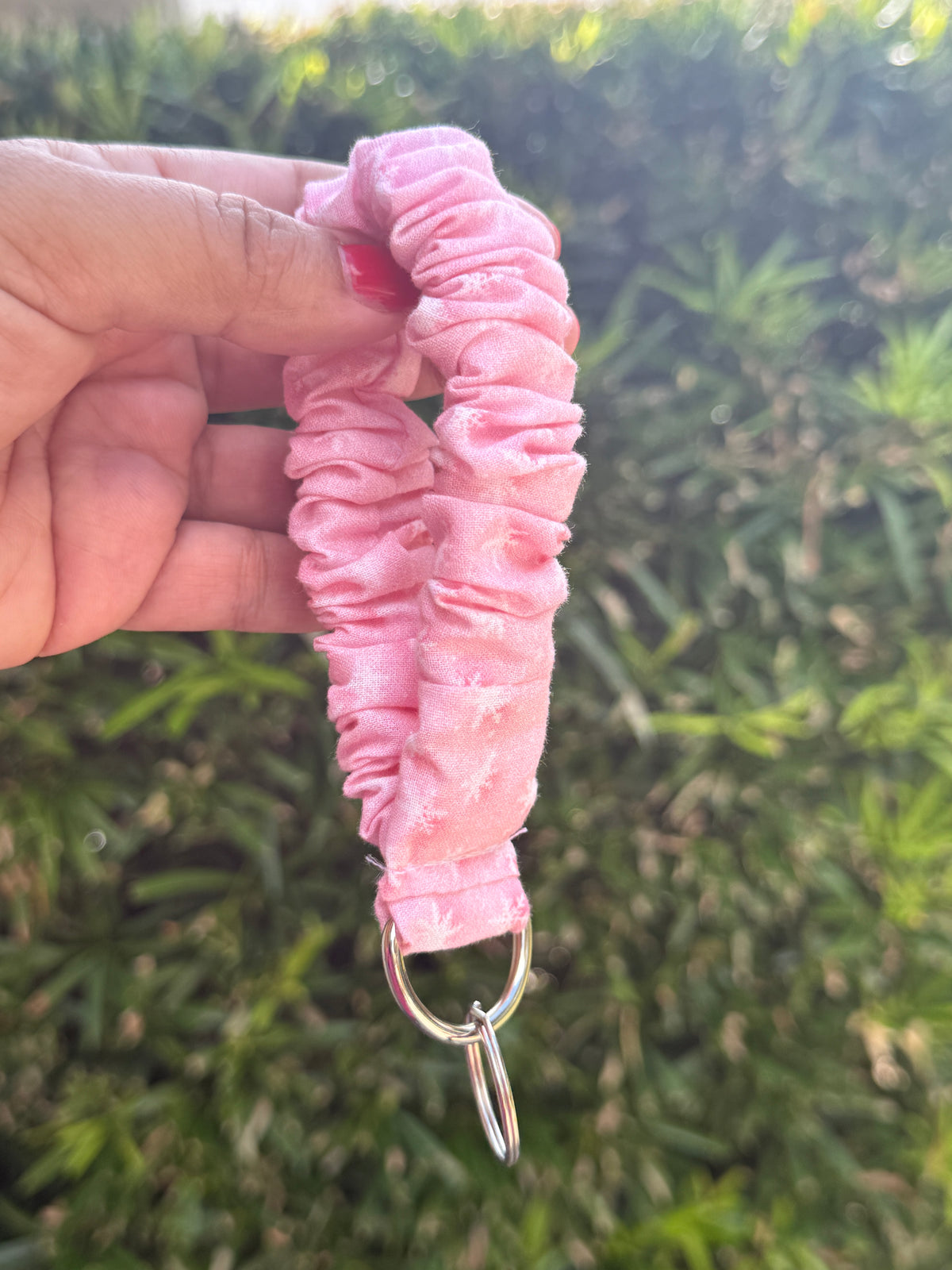 A hand holding a pink ruched fabric wristlet strap with a silver metal D-ring, set against a blurred green outdoor background.
