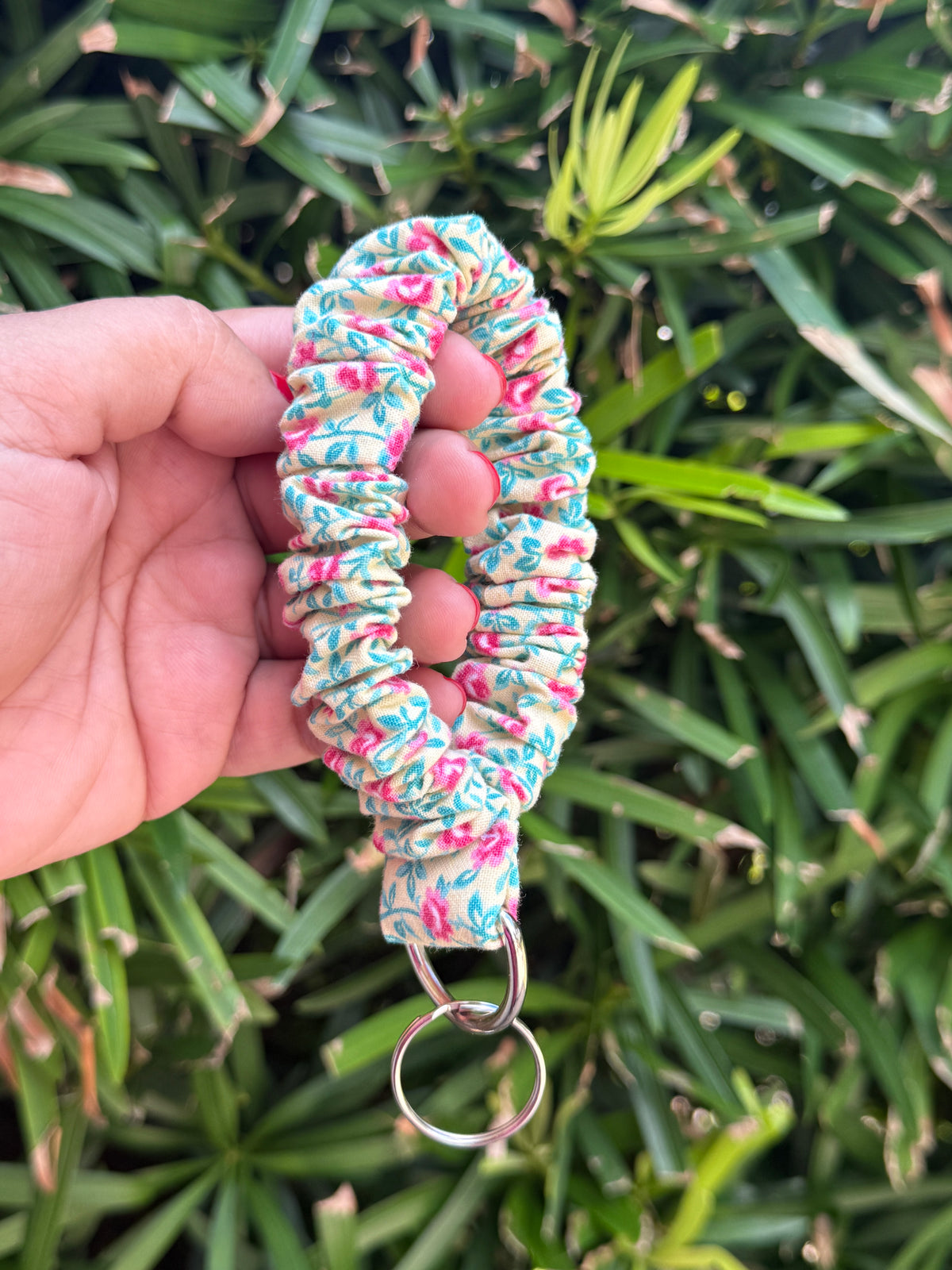 A fabric wristlet keychain with a colorful floral pattern in pink, blue, and green on a cream background, held by a hand against a background of green foliage.